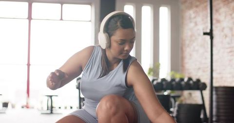 Female athlete squatting and reaching for dumbbell while wearing headphones in sunlit gym interior