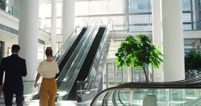 Business professionals walk through a spacious and modern office building lobby with a large escalator. The setting is bright and airy, with contemporary architecture and interior design featuring glass walls and tall pillars. Ideal for use in corporate presentations, business websites, promotional materials, and articles related to professional environments and workplace culture.