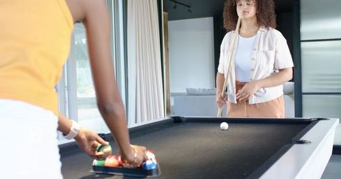 African american women playing pool at home, racking balls and holding cue in sunlit room