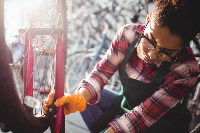 African-American female mechanic is repairing a bicycle in a bike workshop. She is wearing glasses, a plaid shirt, and gloves, focused on tightening a bolt on the wheel. The workshop is filled with various cycling equipment in the background. This image is ideal for promoting cycling services, bike maintenance workshops, or representing skilled trades and craftsmanship.