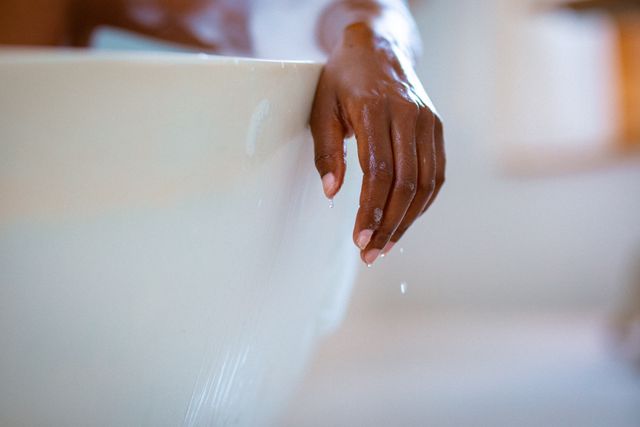 Close-up of an African American woman's hand resting on the edge of a bathtub filled with foam. Water droplets are visible on her hand, emphasizing a moment of relaxation and self-care. Ideal for use in articles or advertisements related to wellness, self-care routines, home spa treatments, and lifestyle content.