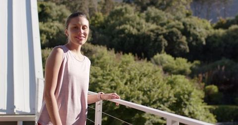Young woman on sunlit balcony wearing smartwatch and pink sleeveless top, smiling