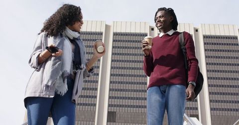 Young diverse coworkers chatting on office steps holding coffee cups and smartphone by office tower