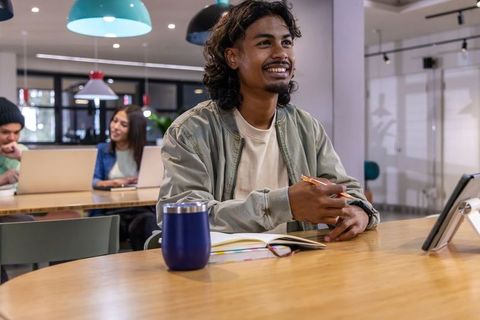Young asian man collaborating in modern coworking space with notebook, laptop and tumbler