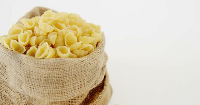 Raw shell pasta filling a burlap sack placed on a white surface and backdrop. Ideal for usage in recipes, cooking blogs, food storage, Italian culinary articles, and kitchen decor promotions.