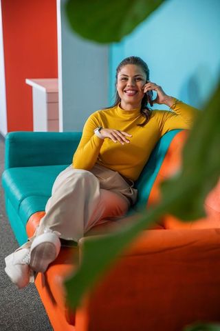 Smiling business woman sitting on colorful sofa talking on smartphone wearing smartwatch