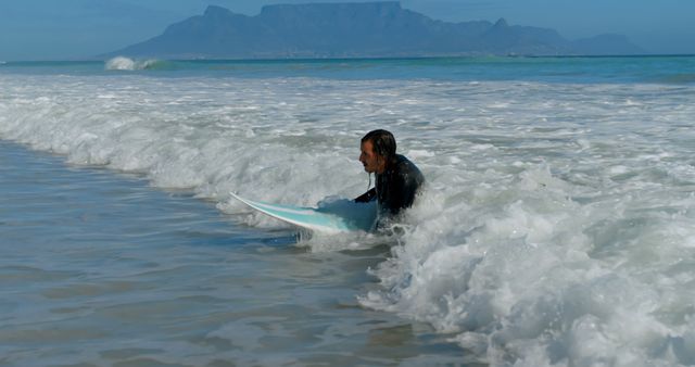 Person seen sitting on surfboard in surf of ocean waves with mountain in background. Ideal for promoting coastal activities, surfing lessons, travel destinations, marine sports, beach vacations, and outdoor adventures.