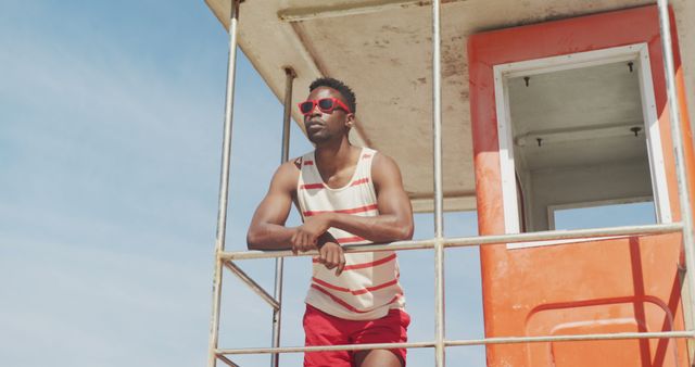 Young man wearing sunglasses and a casual outfit stands on a lifeguard tower overlooking the beach during the day. Great for summer-themed advertisements, lifestyle blogs, travel brochures, and leisure activity promotions.