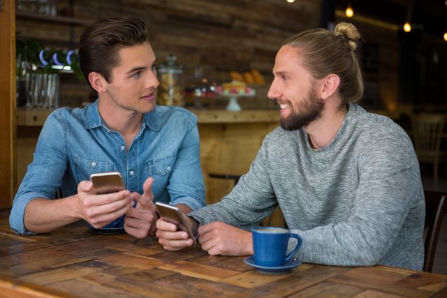 Two young men are sitting at a wooden table in a coffee shop, engaging in a conversation while holding their smartphones. One man is wearing a denim shirt, and the other is in a grey sweater with a man bun. A blue coffee cup is on the table. This image can be used to depict themes of friendship, social interaction, modern technology, and casual meetings in a relaxed setting.