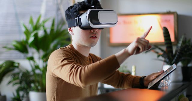 Young man wearing virtual reality headset engaged in a simulation, interacting using hand gestures. Background shows indoor plants and modern home decor creating a comfortable tech-savvy environment. Perfect for illustrating the use of VR technology in everyday home settings, modern gaming, and technological advancements.