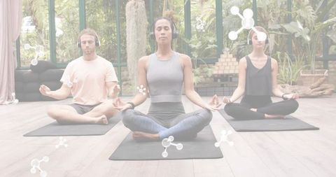 Group meditating with headphones in sunlit glass studio surrounded by lush greenery