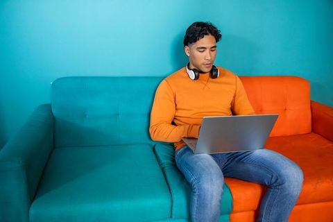 Man working on laptop on teal and orange sofa wearing orange sweatshirt with headphones