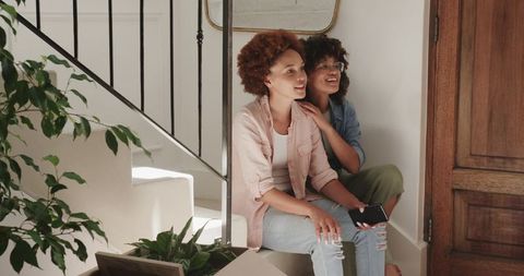 Friends relaxing on stairs holding smartphone and laughing in sunlit modern home