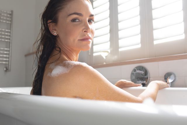 Beautiful woman enjoying a relaxing bubble bath at home, looking serene and peaceful. Ideal for use in articles or advertisements about self-care, wellness, beauty routines, and home spa experiences. Perfect for promoting skincare products, bath accessories, or luxury home amenities.