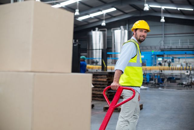 Factory worker in high visibility vest and safety helmet pulling a trolley loaded with cardboard boxes in an industrial warehouse. Ideal for use in articles or advertisements related to manufacturing, logistics, warehouse operations, safety protocols, and manual labor.