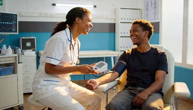 Smiling nurse measuring blood pressure with digital monitor while patient laughing in clinic