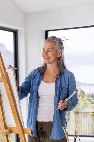 Mature woman painting in home studio by sunlit window holding brushes and smiling in denim shirt