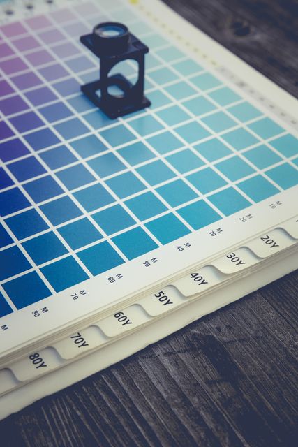 Closeup of a color swatch guide with a magnifying glass used for color matching in printing-related tasks. Ideal for illustrating concepts related to graphic design, print quality control, color accuracy, and design processes in print media.