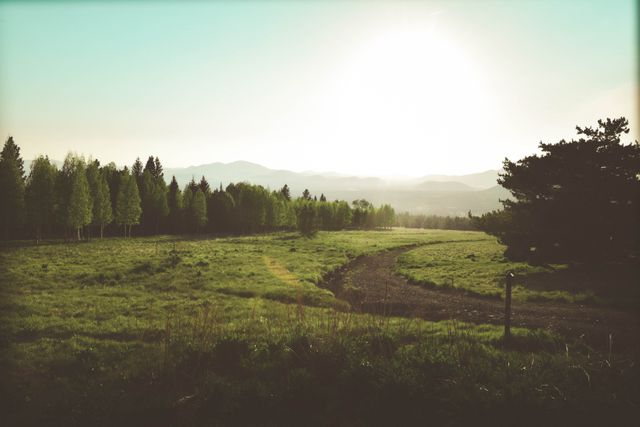 Scenic rural landscape with glowing morning sun rising over distant mountains and green grass fields surrounded by trees. Ideal for environmental campaigns, nature promotions, countryside tourism advertisements, and wallpapers depicting serene outdoor scenes.