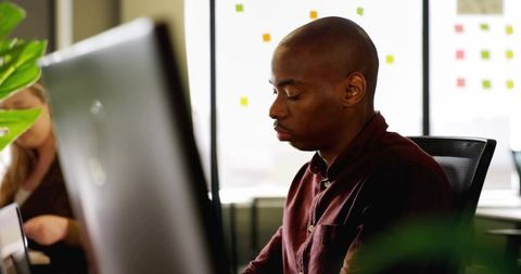 Typing man wearing maroon shirt at modern office desk with sticky-note covered window