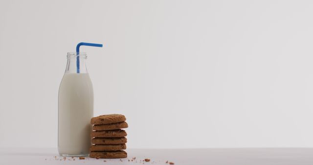 Milk bottle with a blue straw and neatly stacked cookies on a white surface with a white background. Few cookie crumbs around. Useful for ads related to desserts, snacks, beverages, balance diet, baking.