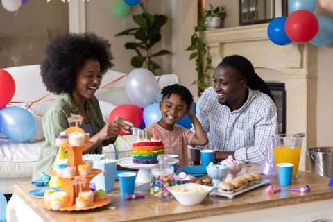 African american family celebrating child birthday cutting rainbow cake with colorful balloons