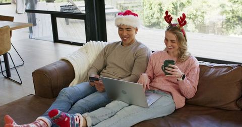 Happy couple shopping online for christmas while lounging on couch with laptop and mug