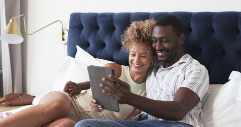 Relaxed diverse couple cuddling on bed streaming tablet in modern navy tufted bedroom