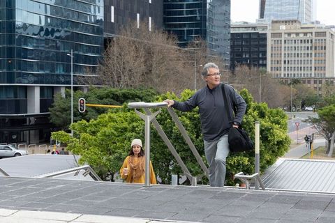 Senior commuters climbing city stairs carrying bag and takeaway coffee near glass towers