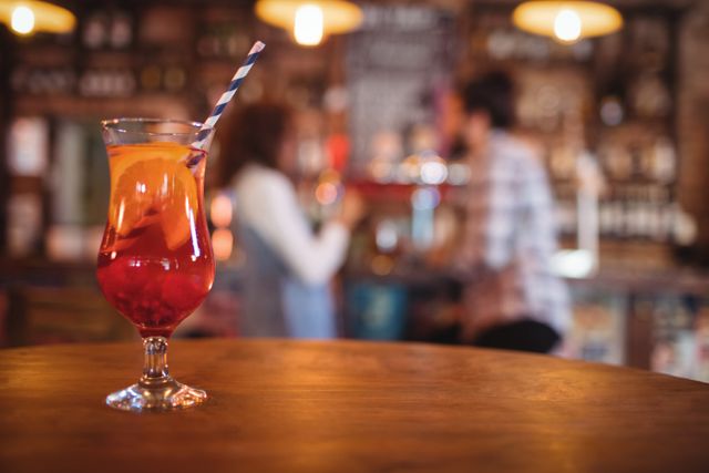 Close-up of a vibrant cocktail drink with a straw and orange slice on a wooden table in a pub. The background shows a blurred scene of people socializing, creating a lively and inviting atmosphere. Ideal for use in advertisements for bars, pubs, nightlife events, or social gatherings.