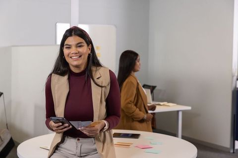Smiling diverse female coworker holding phone and planning at modern office roundtable