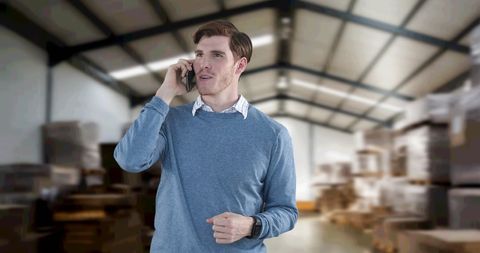 A man is talking on a smartphone inside a spacious warehouse filled with stacked boxes. This image reflects a blend of business communication and logistics, depicting a professional setting's organized environment. The setting can be useful for industries focused on transport, warehousing, supply chain, and business management themes.