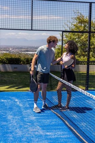 Sunlit diverse couple playing padel on bright blue court with city and mountain views