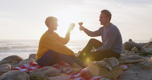 Mature couple enjoying a romantic moment, toasting with wine glasses while picnicking on the beach at sunset. Perfect for illustrating leisure activities, romantic getaways, lifestyle, senior moments, and travel promotions.
