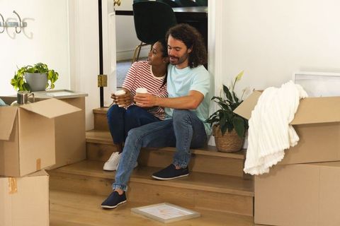 Diverse couple sitting on stairs holding coffee while unpacking boxes after moving house
