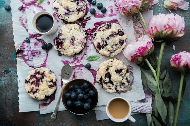 Delicious berry scones arranged with fresh blueberries, cups of coffee, and pink flowers. Ideal for content related to breakfast, baking recipes, homemade snacks, or a cozy breakfast setting. Great for food blogs, social media posts on baking, or advertisements for coffee shops and bakeries.