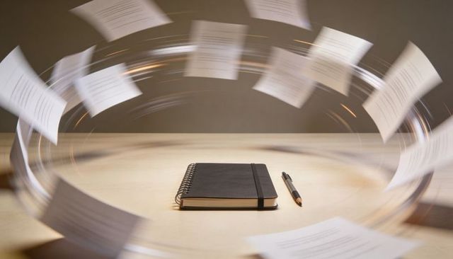 Swirling papers circling black spiral notebook with elastic band and pen on wooden desk