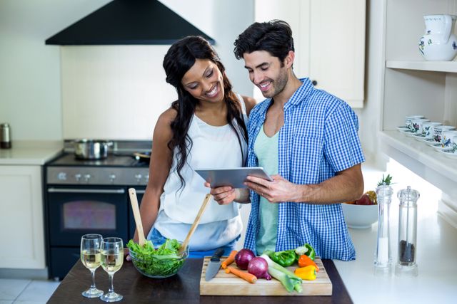 Young couple using digital tablet while preparing a meal in a modern kitchen. They are surrounded by fresh vegetables and salad, indicating a healthy lifestyle. This image can be used for promoting cooking classes, healthy eating, kitchen appliances, or lifestyle blogs.