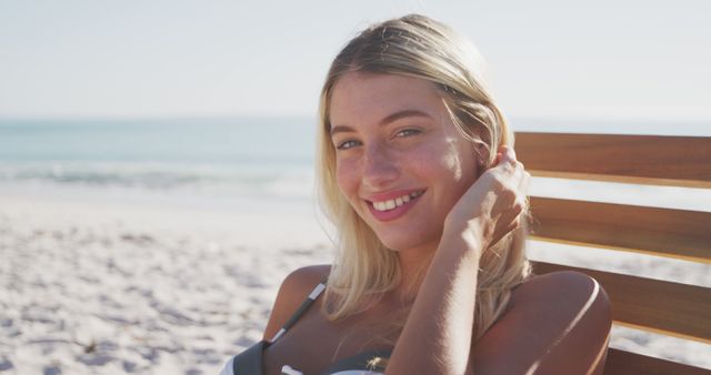 Woman with blond hair smiling while sitting on a chair on the beach. Useful for themes related to summer vacations, beach relaxation, enjoyable outdoors, tropical travel destinations, happiness, and leisure activities.