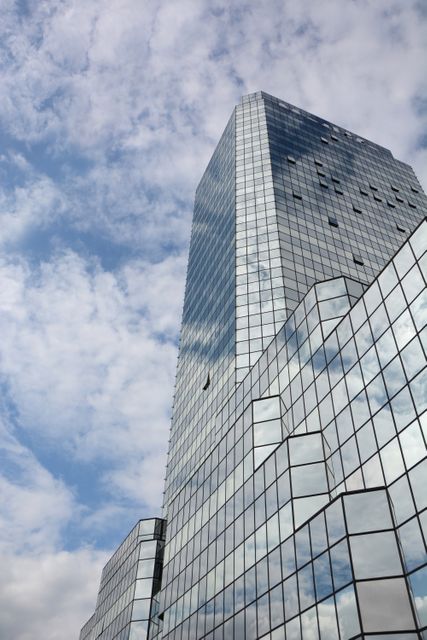 Tall glass skyscraper with reflective windows, set against a blue sky with clouds. Ideal for use in architectural publications, real estate presentations, urban landscape themes, or business and corporate materials to convey modernity and success.
