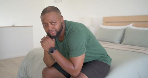 Contemplative man sitting on bed edge leaning forward clasping hands by chin