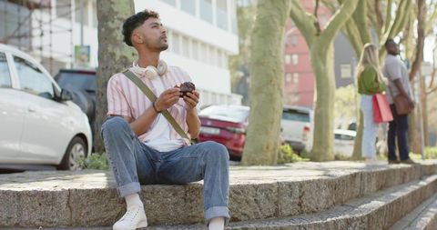 Man sitting on city stone ledge holding camera and wearing headphones, casual urban leisure