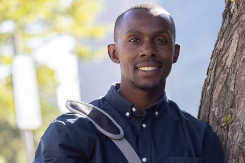 African american student smiling on campus near tree with backpack strap and sunlit bokeh