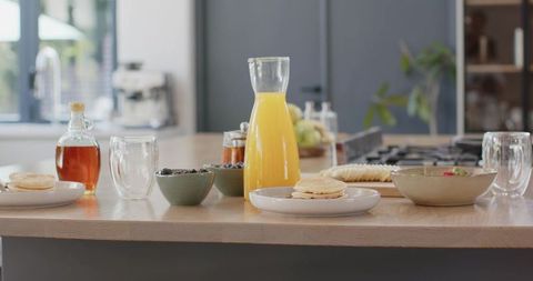 Sunlit kitchen breakfast featuring orange juice carafe, pancakes, berries and cereal