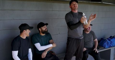 Baseball teammates relaxing on dugout bench sharing water bottle and laughter