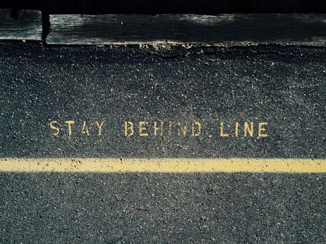 Stay Behind Line warning text on asphalt road with yellow line. Useful for illustrating concepts related to safety, transportation rules, and compliance. Can be used in educational materials, road safety campaigns, and instructional signage design.