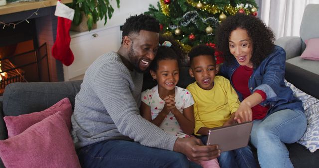 Happy family video calling with relatives while sitting on couch near Christmas tree in cozy living room. Children and parents smiling and enjoying festive season together. Perfect for illustrating family holidays, celebrations, and video call technology.
