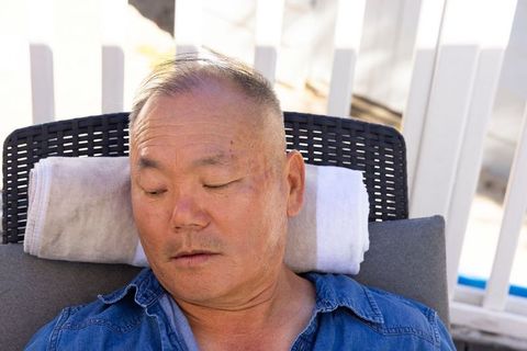 Mature asian man reclining on poolside lounge chair with rolled towel and denim shirt
