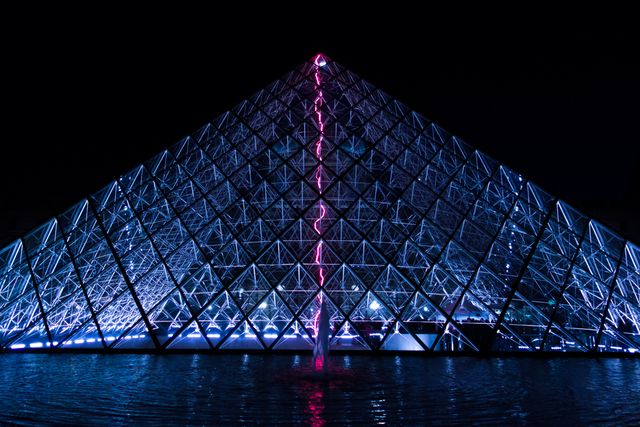 Illuminated glass pyramid structure reflecting in calm water during nighttime. The colorful lights create an appealing visual effect, making it suitable for travel websites, architectural portfolios, and cityscape photography collections. This striking architectural work can enhance materials related to modern design, tourist attractions, and nighttime photography.