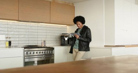 Mid-adult man leaning on white cabinet holding tablet in modern kitchen smiling